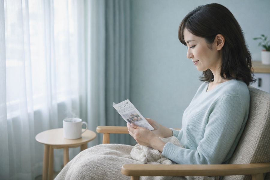 朝の窓辺の木製チェアに座りパンフレットに目を落とす日本人女性の横姿とベージュのブランケット