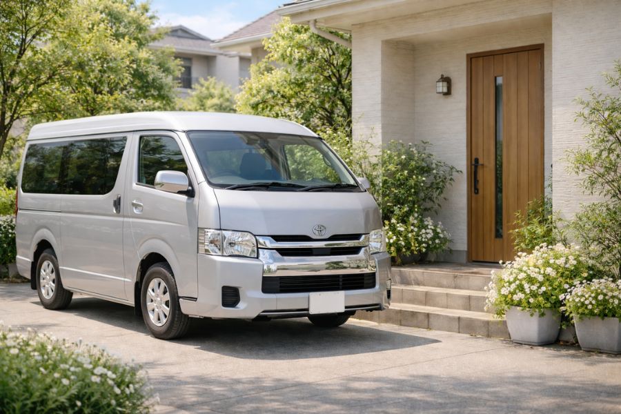 晴れた住宅街の玄関前に停まったシルバーのハイエースと季節の花